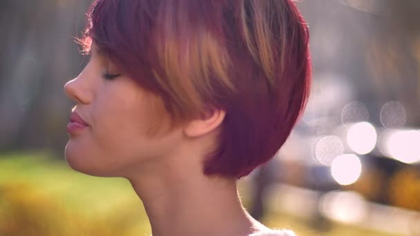 Retrato de cerca de la joven caucásica de pelo rosa girando a la cámara y mirando con perplejidad en el parque otoñal . — Vídeos de Stock