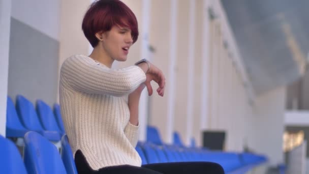 Retrato de la feliz chica de pelo rosa bonita sentada en el estadio y llamando usando su reloj . — Vídeo de stock