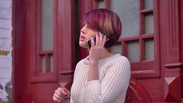 Retrato de jovem caucasiano rosa de cabelos menina atentamente falando no celular no fundo da porta vermelha . — Vídeo de Stock
