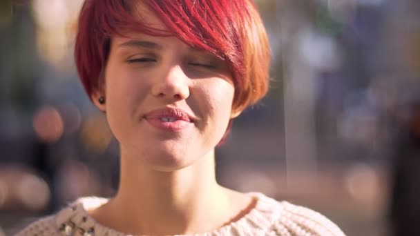 Close-up portrait of young caucasian pink-haired girl smiling humbly into camera on blurred street background. — Stock Video