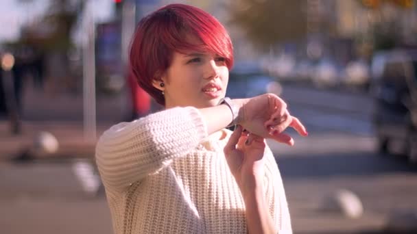 Portrait d'heureuse jolie fille aux cheveux roses appelant en utilisant sa montre sur fond de rue floue . — Video