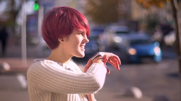 Retrato de chica de pelo rosa bonita emocional llamando usando su reloj en el fondo borroso de la calle . — Vídeo de stock