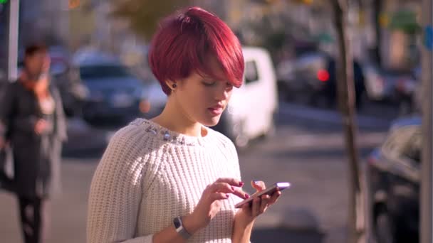 Portrait of pretty pink-haired girl warking with smartphone and turning to camera on blurred street background. — Stock Video