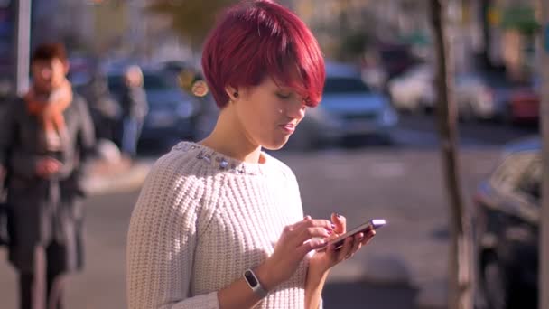 Портрет занятой розововолосой девушки, дергающейся со смартфоном, поворачивающейся к камере и улыбающейся на размытом фоне улицы . — стоковое видео