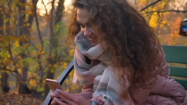 Portrait of caucasian curly-haired woman sitting on bench and making photos using smartphone in autumnal park. — Stock Video
