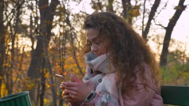 Ritratto di donna caucasica dai capelli ricci seduta sulla panchina e che guarda felicemente nello smartphone nel parco autunnale . — Video Stock