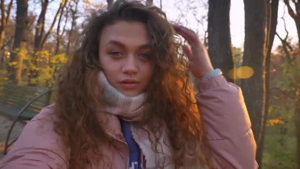 Self-video of caucasian curly-haired woman fixing her hair and making selfie-photos in autumnal park. — Stock Video