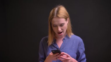 Siyah arka plan üzerine pozitif sürpriz ile akıllı telefon içine izlerken tişörtlü beyaz sarışın kadın portresi.