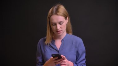 Mutlu bir şekilde smartphone izliyor ve siyah arka plan üzerine gülen tişörtlü beyaz sarışın kadın portresi.