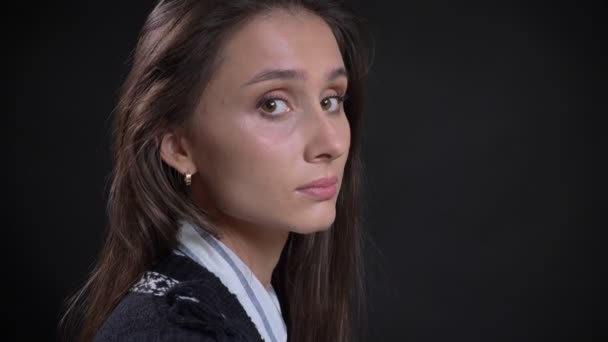 Retrato de una joven caucásica de pelo largo se gira hacia la cámara y observa tranquilamente en ella sobre un fondo negro . — Vídeos de Stock