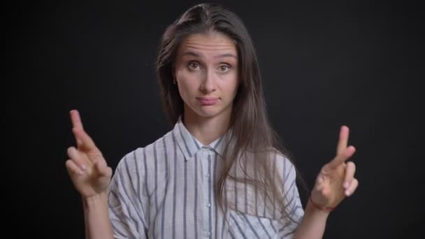 Retrato de una hermosa mujer caucásica de pelo largo haciendo gestos de dedos cruzados para mostrar esperanza de suerte en el fondo negro . — Vídeos de Stock