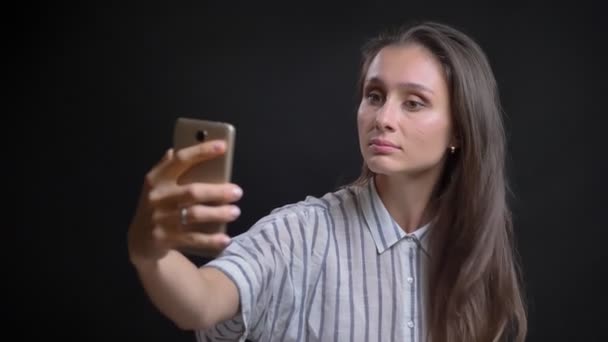 Retrato de una joven mujer caucásica de pelo largo haciendo selfies-fotos usando un teléfono inteligente sobre fondo negro . — Vídeo de stock