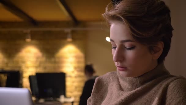 Retrato de cerca de una joven somnolienta de pelo corto que trabaja con una computadora portátil y bosteza en la oficina . — Vídeos de Stock