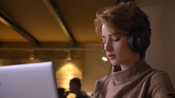 Close-up Portret młodej kobiety krótkowłose, umieszczenie na jej słuchawki i przeniósł się do muzyki, siedząc w biurze. — Wideo stockowe