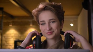 Yakın çekim gülümseyerek onu kulaklıktan koyarak ve ofiste oturan müzik hareketli genç kısa saçlı kadın portresi.