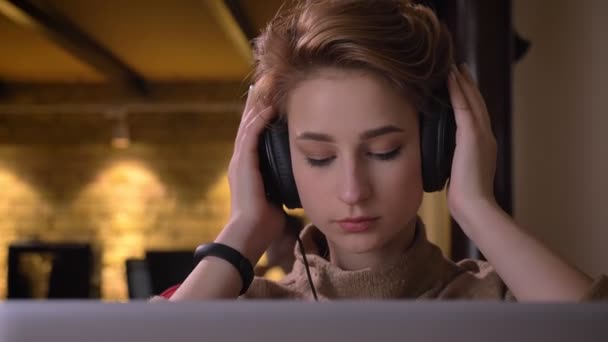 Close-up portrait of young short-haired woman putting on her headphones and slightly moving to music sitting in office. — Stock Video