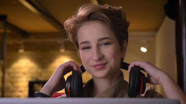 Detailní portrét mladé ženy krátké vlasy s úsměvem položila na její sluchátka a přestěhování do hudba sedí v kanceláři. — Stock video