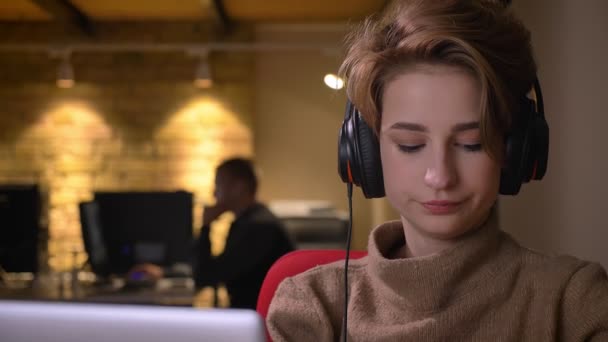 Detailní portrét mladé ženy krátké vlasy ve sluchátkách pozorně pracovat s notebookem a pití nápojů v úřadu. — Stock video