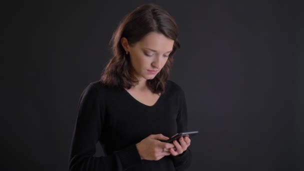 Портрет молодой привлекательной кавказской девушки с длинными волосами, внимательно смотрящей в смартфон на черном фоне . — стоковое видео