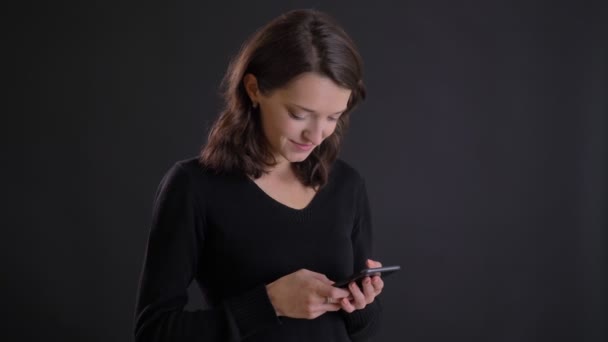 Retrato de la joven atractiva chica de pelo largo caucásica sonriendo mirando en el teléfono inteligente sobre fondo negro . — Vídeo de stock