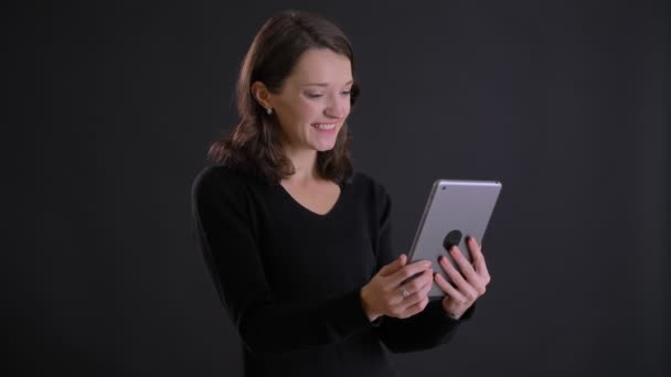 Portrait de jeune fille aux cheveux longs caucasienne attrayante parlant avec sourire en vidéochat à l'aide d'une tablette sur fond noir . — Video