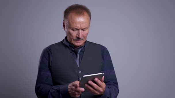 Closeup portrait of concentrated senior caucasian man typing on the tablet with the background isolated on gray — Stock Video