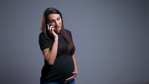 Closeup portrait of pregnant young caucasian female cheerfully talking on the phone and holding her abdomen in front of the camera — Stock Video