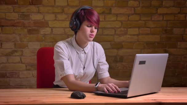 Portrait de jeune blogueuse aux cheveux courts tapant sur ordinateur portable et parlant sur fond de mur de poulet . — Video