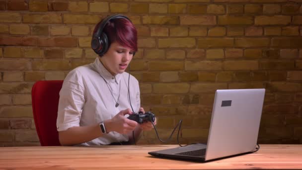 Portrét mladé krátké vlasy ženské návazce sluchátka nervózně hrát pomocí joysticku na bricken zeď na pozadí. — Stock video