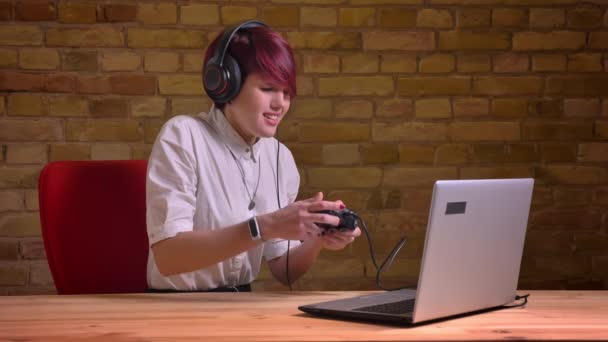 Portrait de jeune femme aux cheveux courts streamer dans les écouteurs jouant joyeusement en utilisant joystick sur fond de mur de poulet . — Video