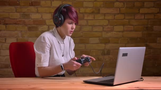 Portrait de femme aux cheveux courts streamer dans un casque jouant nerveusement à l'aide d'un joystick et parlant sur fond de mur de poulet . — Video