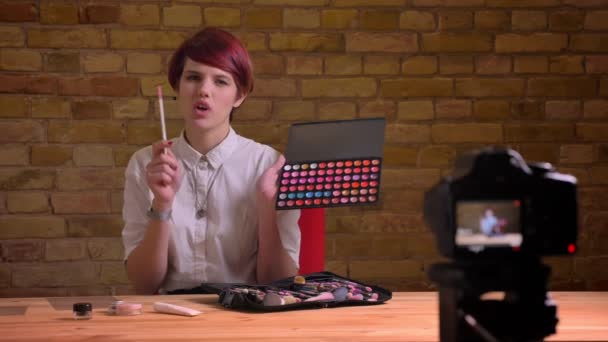 Portrait of young short-haired female beauty-blogger joyfully talking about lipsticks into camera on bricken wall background. — Stock Video