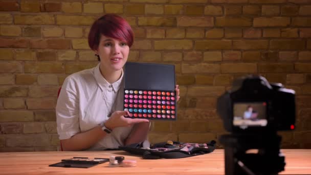 Portrait of young short-haired female beauty-blogger attractively talking about lipsticks into camera on bricken wall background. — Stock Video