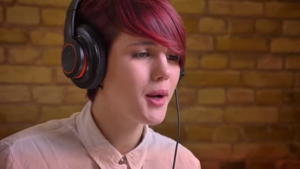 Close-up portrait of short-haired female streamer in headphones talking on bricken wall background. — Stock Video