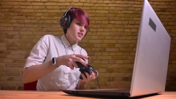 Portrait of young short-haired female streamer in headphones emotionally playing using joystick on bricken wall background. — Stock Video