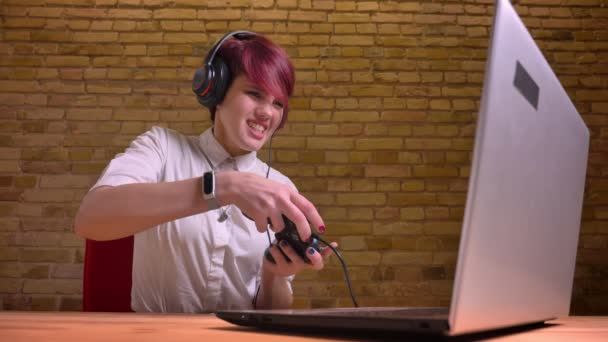 Short-haired girl streamer with manicure and headphones emotionally playing joystick on bricken wall background. — Stock Video