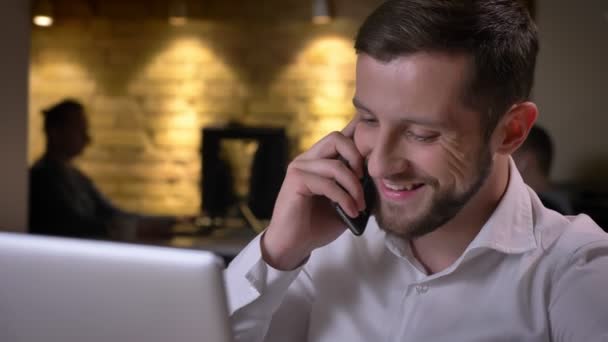 Крупный план молодого веселого кавказского бизнесмена разговаривающего по телефону улыбающегося и смеющегося сидящего перед ноутбуком — стоковое видео