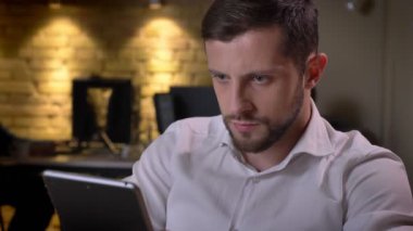 Closeup portrait of young focused caucasian businessman browsing on the tablet in the office on workplace