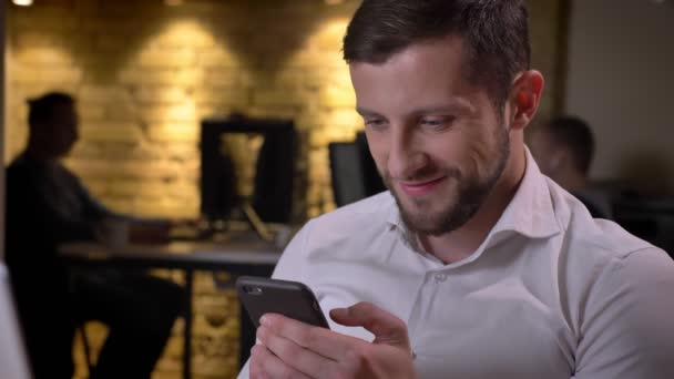 Крупный план молодого веселого кавказского бизнесмена, просматривающего телефон и улыбающегося сидящего перед ноутбуком в офисе — стоковое видео