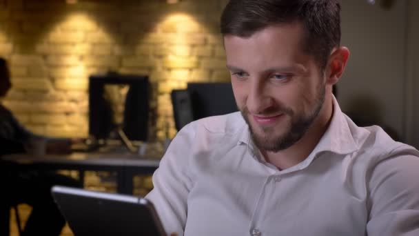 Closeup portrait of young cheerful caucasian businessman typing on the tablet and smiling in the office on workplace — Stock Video