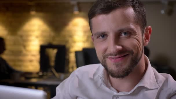 Closeup of happy caucasian businessman sitting in front of the laptop looking at camera and smiling in the office on workplace — Stok video