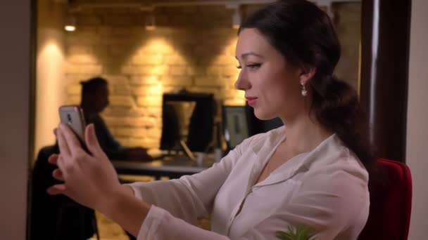 Closeup portrait of young attractive female office worker taking selfies on the phone in front of the laptop on the workplace — Stock video