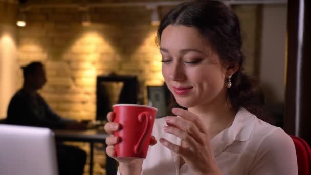 다시 앉아서 웃 고 있는 커피의 컵을 즐기는 직장에 젊은 백인 여성 사무실 근로자의 근접 촬영 초상화 — 비디오