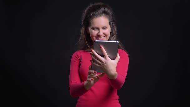 Портрет білої жінки в червоному посміхненому розмові у відеочаті з використанням планшета на чорному тлі . — стокове відео