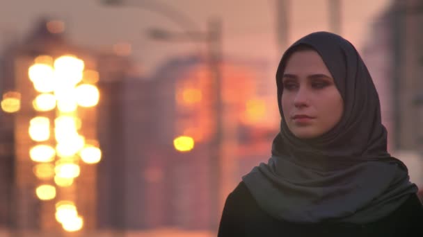 Retrato de cerca de una joven mujer musulmana atractiva en hijab mirando con una sonrisa melancólica a la cámara con la ciudad urbana brillante en la parte posterior — Vídeos de Stock