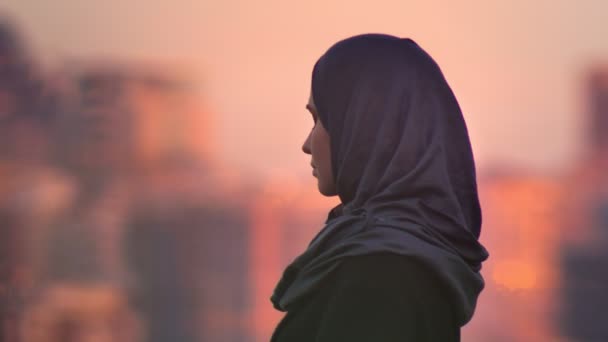 Potret close up dari wanita muda muslim yang menarik melihat ke depan dan beralih ke kamera dengan kota sore yang kabur di bagian belakang — Stok Video