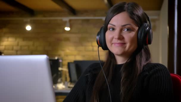 Portret zbliżenie dorosły kaukaski kobieta interesu w słuchawki z przodu laptopa, słuchanie muzyki i patrząc na kamery uśmiechający się — Wideo stockowe