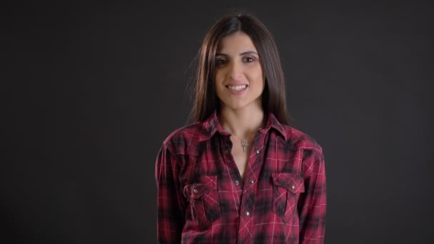 Caucasian long-haired brunette girl with arms crossed modestly smiling into camera and biting her lips on black background. — Stock Video