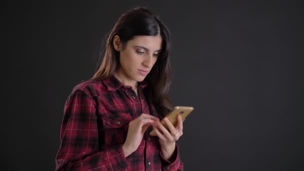 Retrato de joven y hermosa chica de pelo largo caucásica mirando atentamente en el teléfono inteligente sobre fondo negro . — Vídeo de stock