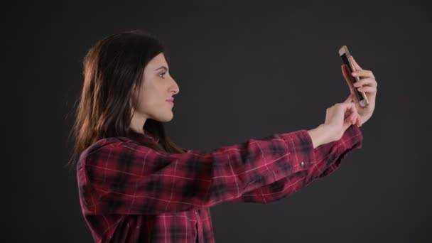 Retrato de joven y hermosa chica de pelo largo caucásica haciendo selfie-fotos usando teléfono inteligente sobre fondo negro . — Vídeo de stock